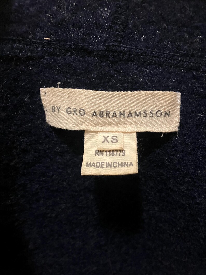 Puede incluir: Primer plano de una etiqueta de prenda azul marino. La etiqueta dice "BY GRO ABRAHAMSSON" y "XS". Debajo de la talla, la etiqueta indica "RN 118779" y "MADE IN CHINA". La prenda parece ser un su&eacute;ter o c&aacute;rdigan.
