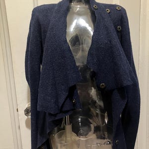 Puede incluir: Un c&aacute;rdigan azul marino con un dise&ntilde;o drapeado &uacute;nico. El su&eacute;ter presenta m&uacute;ltiples cierres de botones y un cuello alto. La prenda se exhibe en un maniqu&iacute; transparente, mostrando su corte asim&eacute;trico y su tejido de punto texturizado.
