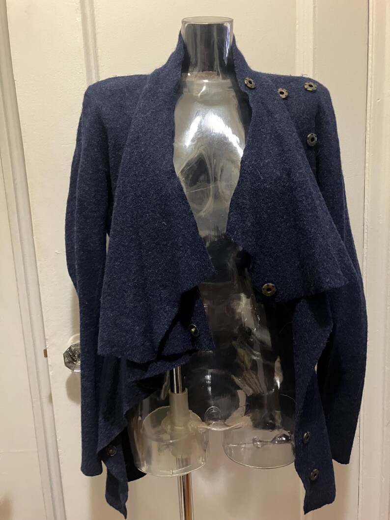 Puede incluir: Un c&aacute;rdigan azul marino con un dise&ntilde;o drapeado &uacute;nico. El su&eacute;ter presenta m&uacute;ltiples cierres de botones y un cuello alto. La prenda se exhibe en un maniqu&iacute; transparente, mostrando su corte asim&eacute;trico y su tejido de punto texturizado.