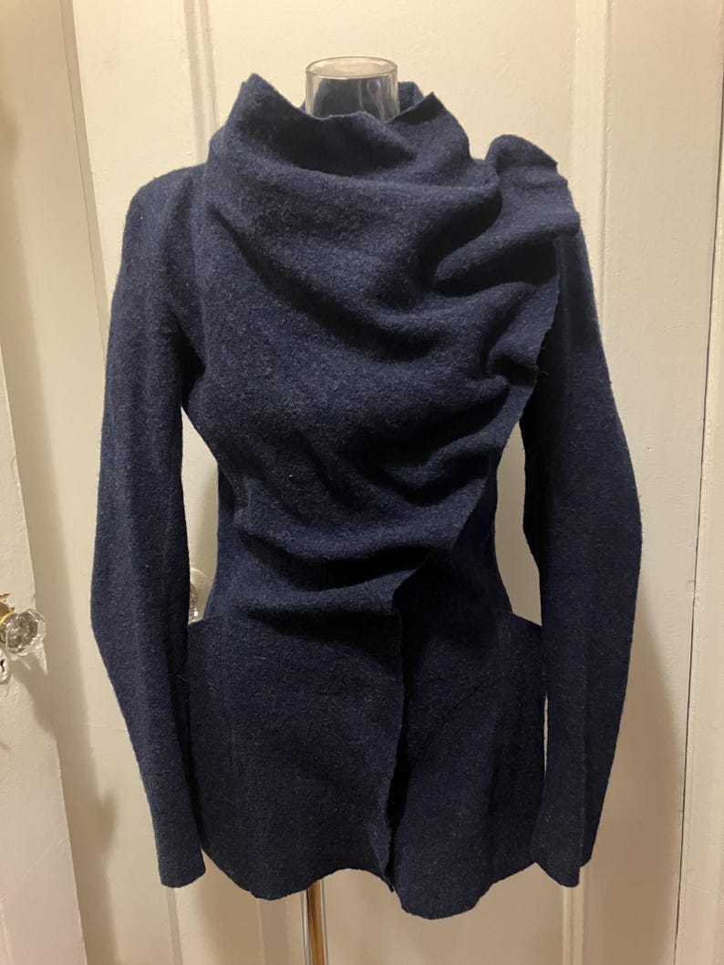 Puede incluir: Una chaqueta de lana azul marino con un cuello drapeado &uacute;nico y mangas largas. La chaqueta tiene un corte entallado y un cierre de bot&oacute;n. La tela parece ser una lana suave y texturizada.