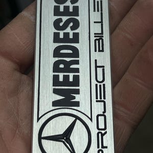 Könnte beinhalten: Ein rechteckiges, gebürstetes Metallabzeichen mit dem Wort "MERDESES" in fetten, schwarzen Großbuchstaben oben. Darunter befindet sich ein schwarzes Mercedes-Benz-Logo und die Worte "PROJECT BILLET" sind vertikal auf der rechten Seite ausgerichtet.