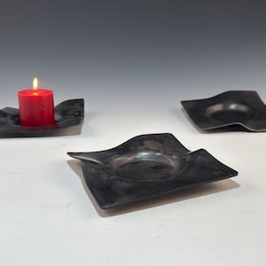 May include: Three black, square-shaped candle holders with a wavy design. One holder contains a lit red candle. The other two are empty. The candle holders are on a white surface.