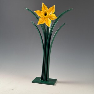 May include: A decorative daffodil sculpture. The flower has a bright yellow, star-shaped bloom with a dark center. The green stem and leaves rise from a rectangular base. The sculpture is made of metal and has a glossy finish.