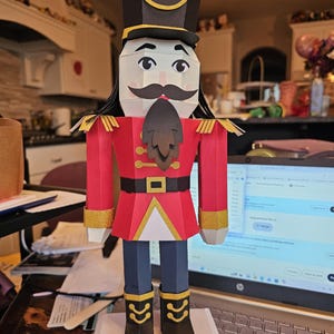 May include: A papercraft nutcracker soldier in a red coat with gold trim, black boots, and a tall black hat with gold accents. The figure has a white face with a black mustache and beard. A festive holiday decoration.