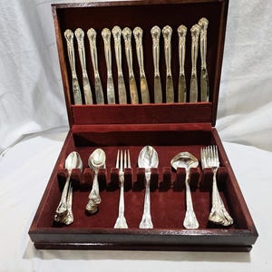 Peut inclure: Un ensemble de couverts argentés disposé dans un coffret de rangement en bois foncé avec un intérieur en velours rouge profond. Le set comprend plusieurs couteaux rangés verticalement dans le couvercle, ainsi que des cuillères, des fourchettes et une louche.