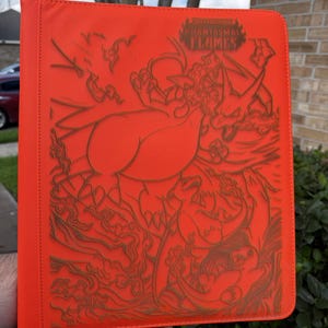 May include: Bright orange binder with a detailed embossed design featuring a dragon-like creature and the text "PHANTASMAL FLAMES". The binder is held open, showcasing its interior.