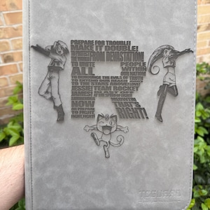 May include: Gray trading card binder with embossed Team Rocket characters and text. The text includes phrases like "Prepare for Trouble!" and "Meowth, that's right!" The binder has a textured surface and a "TeeGuard" logo.