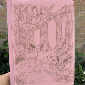 May include: Pink binder with a forest scene featuring engraved images of cartoon characters. The cover has a stitched border and the word "ZOELY" is visible at the bottom. The binder is held by a person.