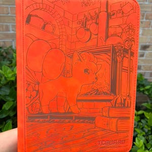 May include: A bright orange card binder featuring an embossed illustration of a Vulpix character standing in front of a fireplace. The design includes kitchenware hanging on a brick wall, books, and the text TCGUARD printed in the bottom corner.