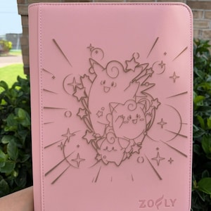 May include: Pink binder with a debossed design featuring cartoon characters and stars. The binder has a stitched edge and the word "ZOFY" at the bottom. The binder is held in front of a brick wall and green foliage.