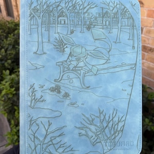 May include: A light blue trading card binder with an embossed illustration of a cartoon character sitting on a park bench in a winter scene. The binder has the text "TOGUARD" and "guard your trading cards" at the bottom.