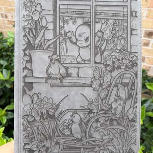 May include: Gray tablet case with an engraved garden scene. The design features birds, flowers, and a window with a cartoon character. The case is rectangular and has a smooth texture.