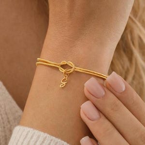 May include: A gold-colored bracelet with a knot design and a small charm with the letter "e". The bracelet is worn on a wrist and has a delicate, elegant appearance. The bracelet is made of a thin, flexible material.