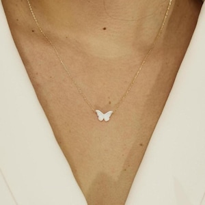 May include: A delicate gold necklace featuring a small, white butterfly pendant. The butterfly is centered on a fine gold chain, creating a minimalist and elegant design. The necklace is shown against a neutral background.