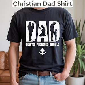 Op de afbeelding: Zwart T-shirt met de tekst "Christian Dad Shirt" bovenaan. Het woord "DAD" staat in grote witte letters met silhouetten van een vader en kind. Onder het woord "DAD" staan de woorden "DEVOTED ANCHORED DISCIPLE" en een kruis-ankersymbool.