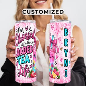 May include: Two cylindrical tumblers with lids and straws featuring a pink leopard print background. The left tumbler displays the text I'm Obsessed with the LOADED TEA Life. The right tumbler features a pink bow, fruit graphics, and the name BRYNI.