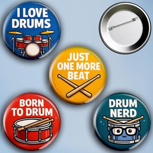 Może przedstawiać: Zestaw pięciu okrągłych przypinek z motywami perkusyjnymi. Przypinki zawierają hasła takie jak "I Love Drums", "Just One More Beat" i "Drum Nerd", a także ilustracje zestawów perkusyjnych i pałeczek.