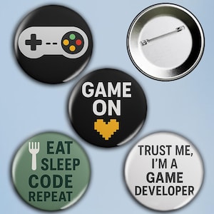 Puede incluir: Un conjunto de cinco botones redondos. Un botón negro muestra un controlador blanco. Otros botones tienen las frases "GAME ON", "EAT SLEEP CODE REPEAT" y "TRUST ME, I'M A GAME DEVELOPER".
