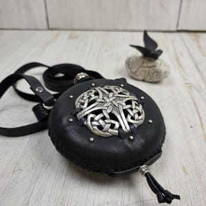 May include: A round black leather pouch featuring a central silver-toned metal emblem with an intricate interwoven knot pattern. The pouch is secured with a black leather strap and includes small silver metal studs and a dangling leather cord.