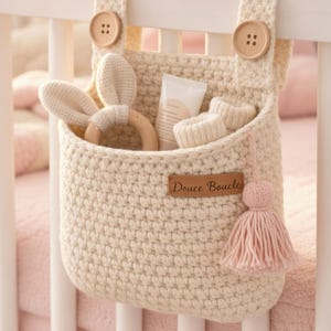 Peut inclure: Organisateur de berceau pour bébé en crochet crème, avec un anneau de dentition en bois en forme de lapin, de petites bottines crème et un tube de lotion. L'organisateur a un pompon rose et est fixé au berceau avec des boutons en bois. L'étiquette indique "Douce Boucle."