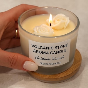 Op de afbeelding: Een brandende Volcanic Stone Aroma Kaars in een heldere glazen pot, met twee witte roosvormige wasdecoraties. De kaars staat op een houten onderzetter. Het label luidt "Christmas Warmth" en "StoneandScentCo".