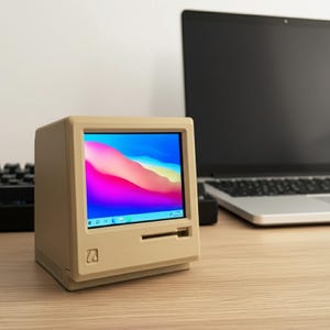 May include: A beige, compact, retro-style computer monitor featuring a square screen displaying a vibrant, abstract, multi-colored wave graphic. The device has a classic boxy frame with a horizontal slot below the screen and a small square logo.