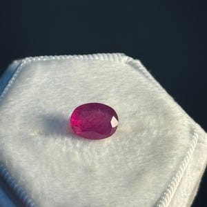 May include: A vibrant, oval-cut ruby gemstone with a deep pinkish-red hue and multifaceted surface, resting centred on a white, hexagonal, velvet-textured display cushion. The gemstone exhibits internal clarity and light reflection.