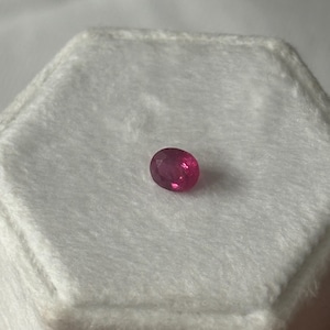 Könnte beinhalten: Ein kleiner, oval geschliffener Edelstein mit einem leuchtenden, tief rosa-roten Farbton. Der Stein weist mehrere Facetten auf, die das Licht reflektieren und für ein funkelndes Aussehen sorgen. Er liegt mittig auf einer weißen, strukturierten Fläche.