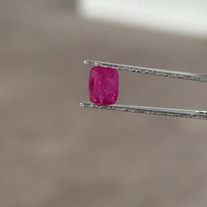 Peut inclure: Une petite pierre précieuse rouge rosé vif avec une taille coussin, maintenue fermement entre les deux fines pointes métalliques d'une pince argentée. La pierre présente une qualité translucide avec des facettes internes visibles.