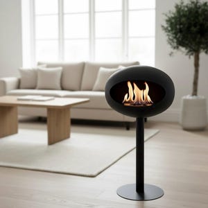 Puede incluir: Una chimenea negra moderna e independiente con una llama visible a través de una abertura ovalada. La chimenea está montada sobre un soporte negro con una base circular. El fondo muestra una sala de estar con un sofá, una mesa de centro y una planta en maceta.