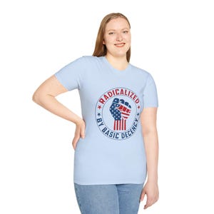 Puede incluir: Camiseta azul claro con un gráfico circular que dice "RADICALIZED BY BASIC DECENCY". El gráfico incluye un diseño de puño en alto con los colores de la bandera americana. La camiseta la lleva una persona.