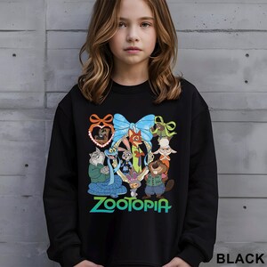 May include: Black sweatshirt featuring a colorful graphic of characters from the animated film "Zootopia". The design includes a blue bow, a heart-shaped frame, and the word "Zootopia" in green and blue. The word "BLACK" is printed at the bottom right.