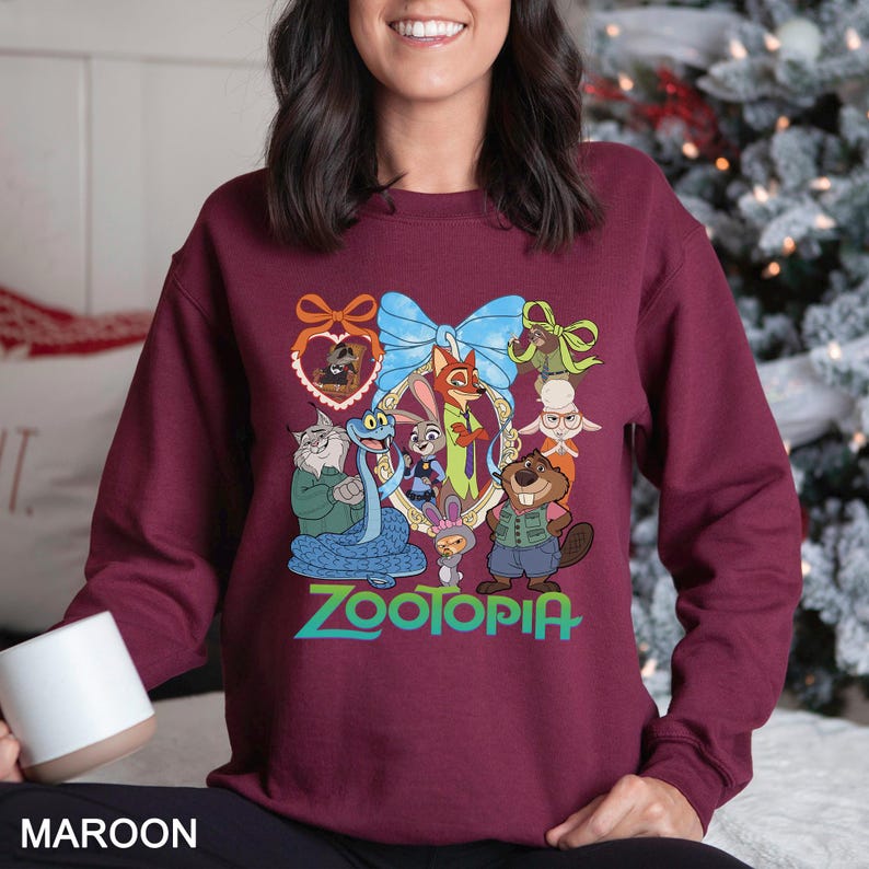 May include: Maroon sweatshirt featuring a colorful graphic of characters from the movie Zootopia. The design includes a blue bow and the word "Zootopia" in green. The sweatshirt is a solid maroon color.