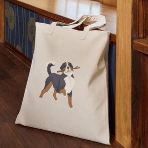 May include: A cream-colored tote bag featuring a Bernese Mountain Dog illustration. The dog is depicted with black, brown, and white markings, holding a stick in its mouth. The bag has two handles and is made of a sturdy fabric.