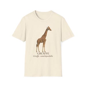 May include: Cream-colored t-shirt featuring a detailed illustration of a giraffe. The giraffe has a brown and tan spotted pattern. Below the giraffe, the word "GIRAFFE" is printed, along with the scientific name, "Giraffa camelopardalis."