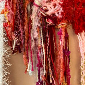 May include: Close-up of a handmade textile with a variety of textures and colors. The piece features crochet work in shades of red, pink, and white, with dangling strands of yarn and embellishments. The yarn strands include metallic threads and small floral accents.