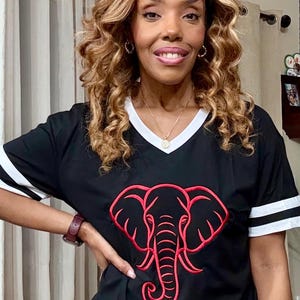 May include: Black V-neck shirt with white stripes on the sleeves and collar. The shirt features a red elephant design on the front. The shirt is made of a soft material.