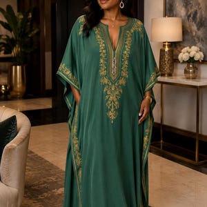 May include: A floor-length dark green kaftan dress featuring gold embroidery along the V-neckline, shoulders, and side panels. The center of the chest displays the letters AKA in a vertical gold font. The garment has wide, flowing sleeves.