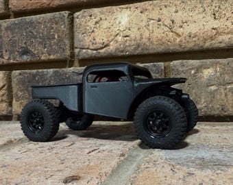 May include: A black toy truck with large, knobby tires. The truck has a detailed body, including a bed, cab, and fenders. The vehicle is set against a brick background.