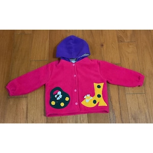 May include: A pink fleece hooded jacket with a purple hood and button closure. The jacket features appliqued designs of a black and green shape and a yellow shape with polka dots. The jacket is displayed on a wooden surface.