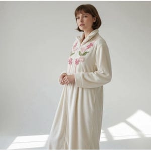 May include: Cream-colored robe with a floral embroidery design in pink and green. The robe has a zip-up front, long sleeves, and a shawl collar. The embroidery is centered on the chest area. The robe is made of a soft, plush material.