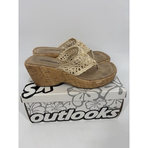 Puede incluir: Un par de sandalias de cuña beige con correas tejidas y suelas de corcho. Las sandalias se exhiben en una caja blanca con el nombre de la marca "outlooks". Diseño de estilo chancla.