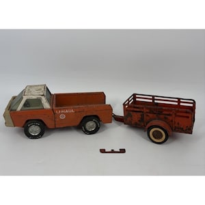May include: Vintage orange and white U-Haul toy truck with a matching trailer. The truck has black tires and the U-Haul logo on the side. The trailer is a simple open-top design. A small metal piece is detached.
