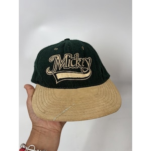 May include: A dark green baseball cap with a tan brim. The word "Mickey" is embroidered in a cursive font with a gold outline. The cap is a classic style, suitable for casual wear.