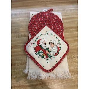 May include: A cream-colored kitchen towel with fringe at the bottom, topped with a quilted potholder. The potholder features a red floral pattern and a central illustration of Santa Claus and Mrs. Claus surrounded by a green holly wreath.