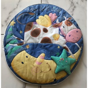 Puede incluir: Una alfombra de juego redonda y acolchada que representa una vaca saltando sobre la luna, con una estrella sonriente. La alfombra tiene un fondo azul con estrellas y el texto "Hey diddle diddle". La vaca es blanca con manchas marrones y orejas rosas.