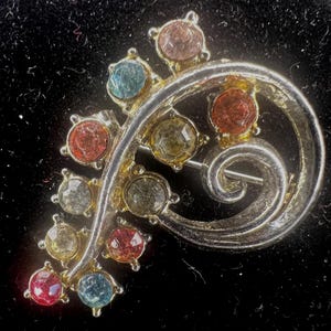 May include: A vintage brooch with a silver-tone metal base and a swirling design. The brooch is adorned with colorful, faceted gemstones in shades of red, blue, yellow, and pink. The pin is set against a black background.