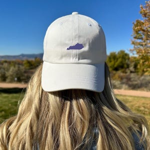 May include: A white baseball cap with a curved brim. The cap features a purple embroidered outline of the state of Kentucky on the front. The hat is worn by a person with long blonde hair.