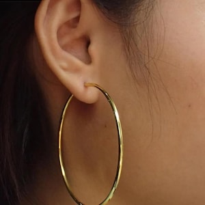 May include: A large, polished gold hoop earring with a thin, circular wire design. The earring is worn in the earlobe, featuring a smooth, reflective metallic surface that catches the light, creating a simple and continuous circular shape.