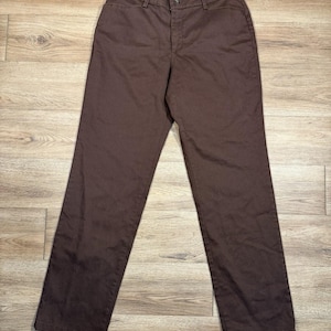 May include: A pair of brown pants with a button and zipper closure. The pants have a classic, straight-leg design and are made of a solid, dark brown fabric. The pants are laid flat on a wooden surface.
